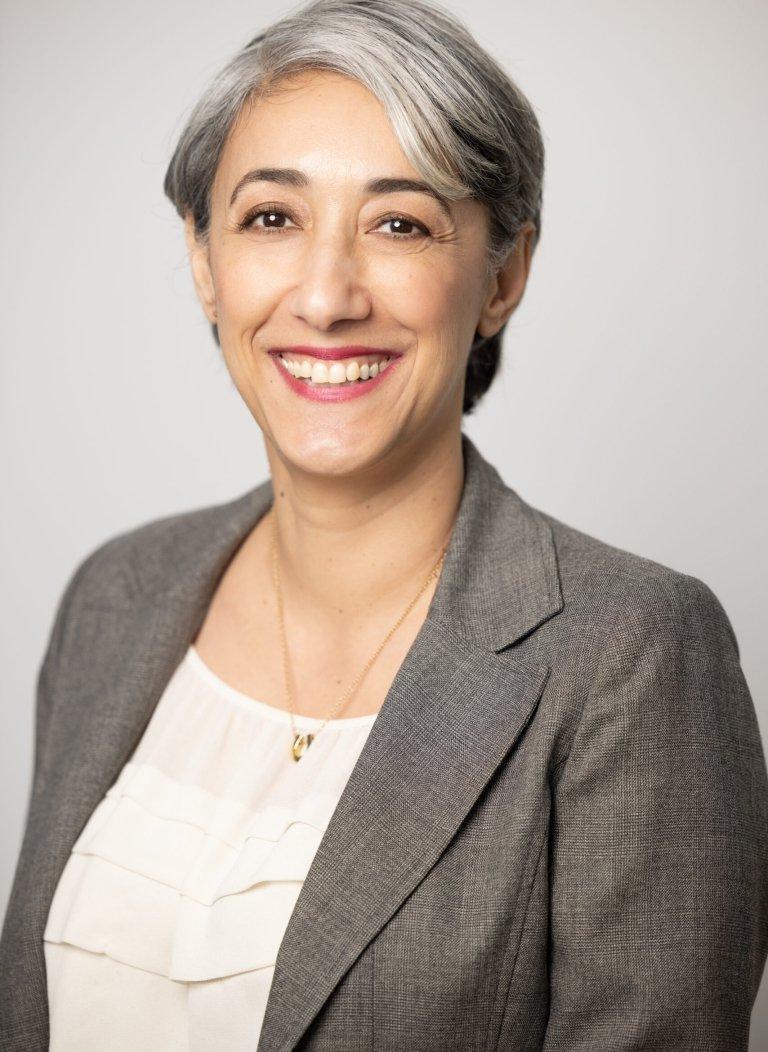 Person with short gray hair, smiling, wearing gray blazer and white shirt, in front of gray background.