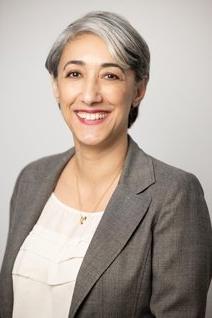 Person with short gray hair, smiling, wearing gray blazer and white shirt, in front of gray background.